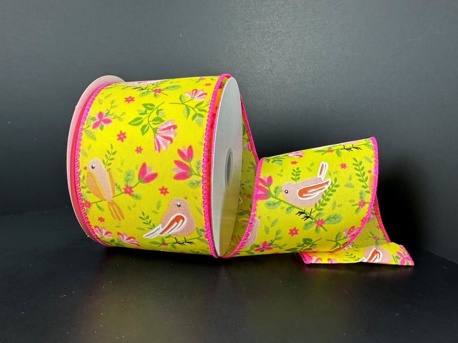 A slightly unrolled 2.5X10Y Yellow Peach Pink Satin Spring Birds and Flowers Ribbon (41542-40-22) sits on a dark surface, displaying its vibrant pattern adorned with birds and flowers on pink-edged satin.