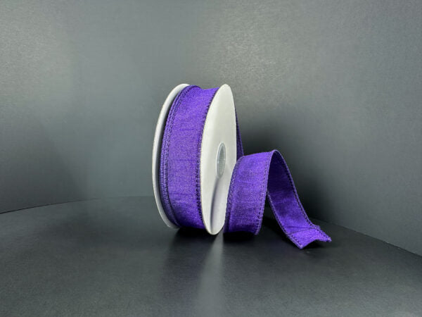 A spool of 7/8X10Y Purple Faux Dupioni Ribbon (72434-05-11) is partially unwound and displayed on a plain dark gray background, ideal for craft ribbon projects.