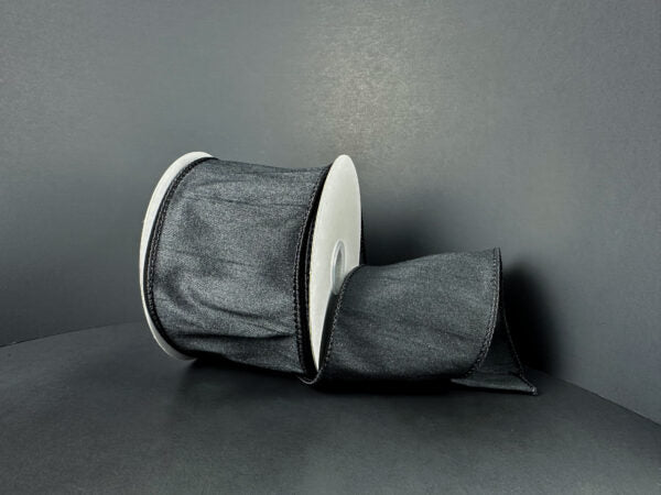 A roll of 2.5X10Y Black Faux Dupioni Ribbon (72434-40-21) with a slightly shiny texture is unspooled and resting on a smooth, dark gray surface against a matching gray background.