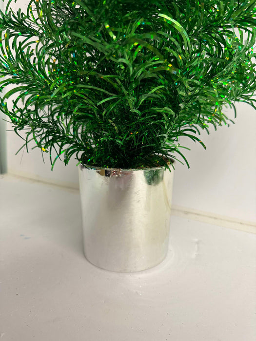 The 24 In Tinsel Tree 84578-Gn, a small artificial green tinsel tree with shiny leaves, sits in a reflective silver pot on a white surface against a white background.