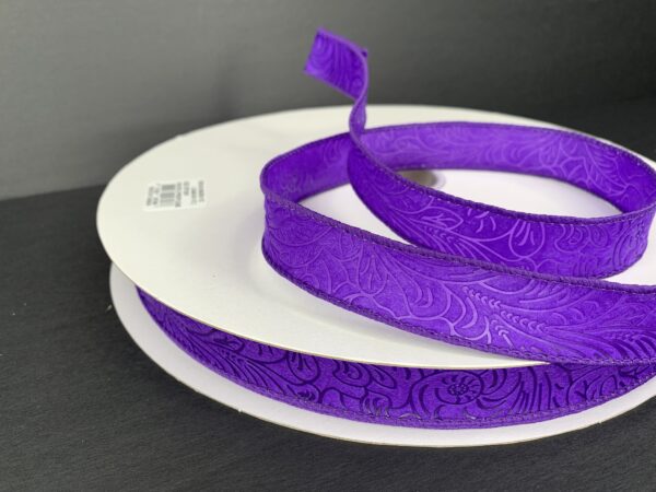 The 7/8X50Y (42466-05-11C) Purple Flower Embossed Ribbon, partially unwound, is displayed on a dark background and elegantly wound around a white circular base.