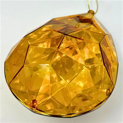 A 6.3" x 4" x 9" Amber Plastic Drop Ornament (DCORN5408AM) with a faceted, geometric design hangs from a thin gold string, reflecting light against a plain white background.