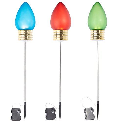 A set of 29" Lighted Frosted Christmas Light Picks, each featuring a metallic base and frosted bulb reminiscent of oversized Christmas lights, glows in blue, red, and green. Mounted on thin metal rods with black ground stakes, these solar-powered garden stake lights are efficiently illuminated by small black solar panels.