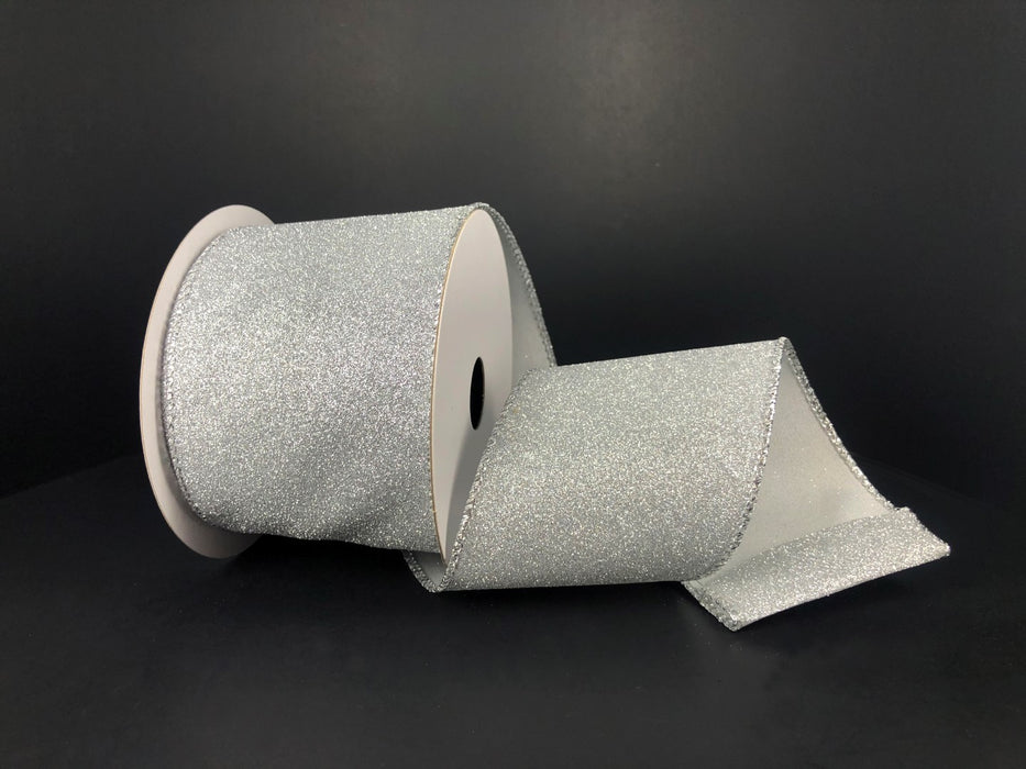 A 2.5X10Y Silver All Flat Glitter Ribbon X820640-16 with a white reel rests on a black surface, partially unrolled to display its sparkling flat glitter texture and finished edges.