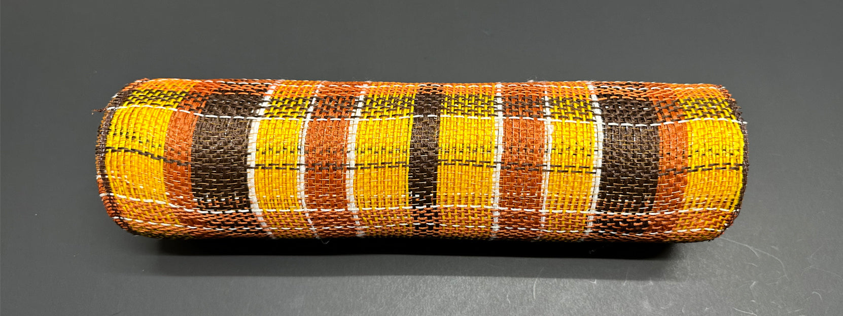 The 10X10Y Rust-Yellow-Chocolate Fall Plaid Mesh XB235310-49, a cylindrical woven object in rust yellow, orange, brown, and white plaid, is placed on a dark surface.