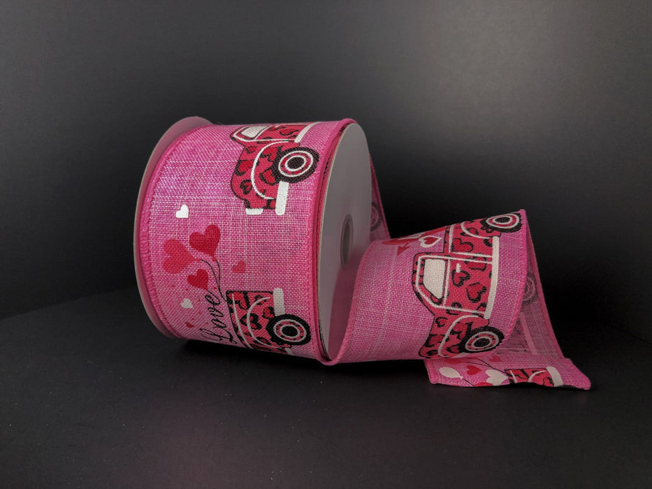 A spool of 2.5x10Y Pink Linen/Pink Cheetah Truck-Hearts ribbon (11265-40-44) featuring pink trucks, hearts, and love designs on a black background is partially unrolled and lies flat in front of the spool.
