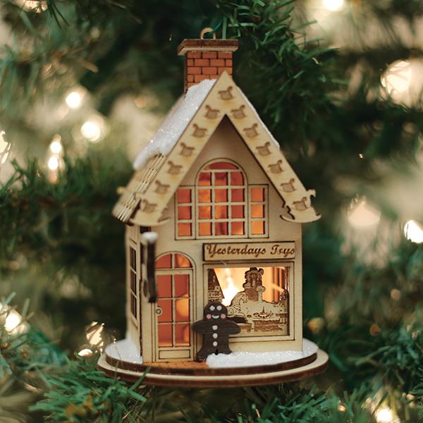 The Yesterday's Toys Old World Christmas Ornament 80004 is a miniature wooden house, warmly lit from inside and dusted with snow, capturing Ginger Cottage charm as it hangs on a Christmas tree amid softly blurred background lights.