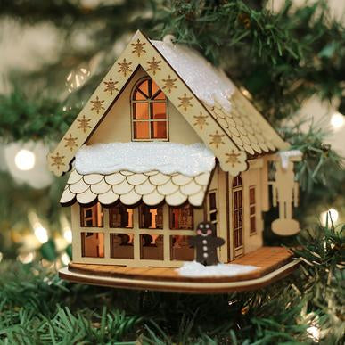 The Drosselmeyer's Nutcracker Old World Christmas Ginger Cottage Ornament 80007, adorned with artificial snow and cutout details, hangs on a lit Christmas tree, featuring a gingerbread figure at the front for a festive touch.