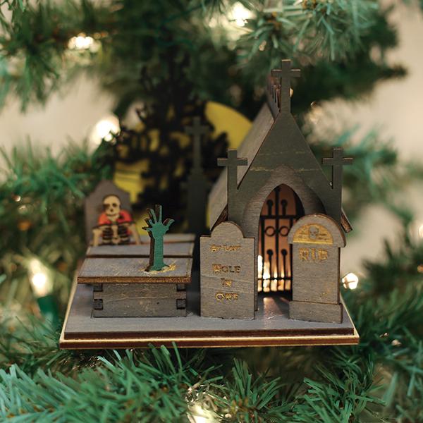 A close-up of the Creepy Cemetery Old World Christmas Ornament 82003 on a tree, showcasing a miniature graveyard with tombstones, a crypt, a skeleton, and a green hand rising from the ground.