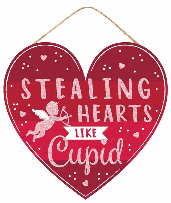 The AP8985 is a 12L x 11.5H heart-shaped Cupid Glitter Sign featuring a Cupid illustration and the phrase Stealing Hearts Like Cupid on a red background with white hearts and dots, complete with an attached rope for easy hanging.