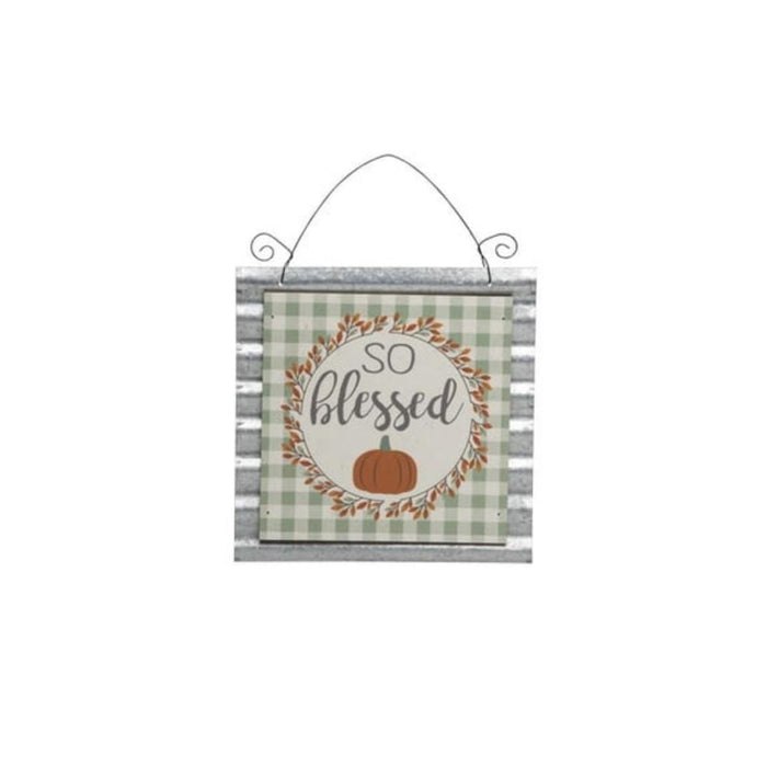 The Small Metal & MDF Thanksgiving Sign Assorted J9365 features a green plaid border, leaf wreath, orange pumpkin, and So blessed in cursive. Hanging by a wire, its perfect fall decor for giving thanks.