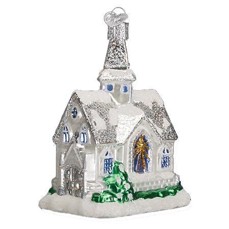 The Sparkling Cathedral 20076 Old World Christmas Ornament is a glass piece shaped like a white church with a glittery silver roof, blue window accents, gold and blue stained glass, green shrubs, and snow at the base.
