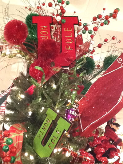 A Christmas tree adorned with red and green ornaments, twinkling lights, red bows, and the RAZ 7" Letter U North Pole Red Green Christmas Ornament Set of 2 creates a festive North Pole holiday theme.