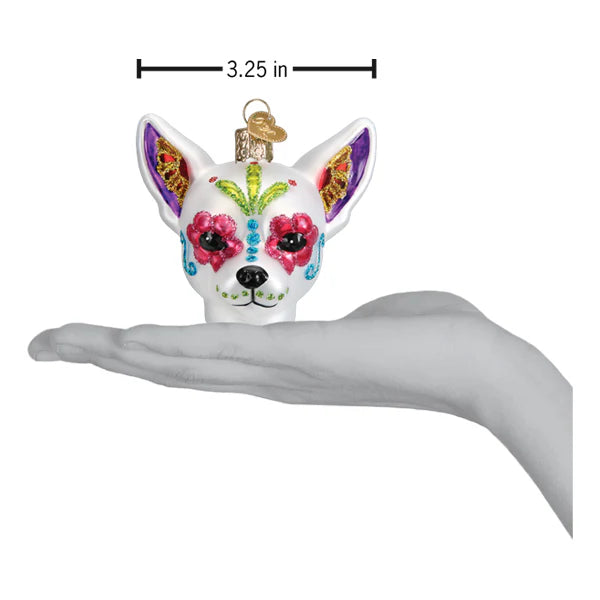 A hand holds the Dia De Los Muertos Dog Ornament Old World Christmas 26097, a 3.25-inch decorative piece shaped like a Chihuahua head and adorned with vibrant Day of the Dead designs in pink, blue, green, purple, and gold—an eye-catching ornament perfect for celebrating the Mexican holiday.