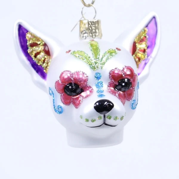 The Dia De Los Muertos Dog Ornament Old World Christmas 26097 is a white dog ornament adorned with colorful, glittery designs in a Day of the Dead style, featuring pink flowers, blue swirls, and gold accents on its face and ears. This unique Mexican holiday ornament captures the festive spirit and vibrant artistry celebrated by Old World Christmas.