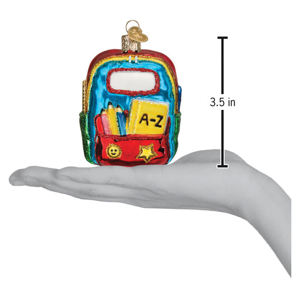 A hand holds a 3.5-inch First Day of School Old World Christmas Ornament 32655, a glass decoration featuring a yellow book labeled A-Z, pencils, and stars.