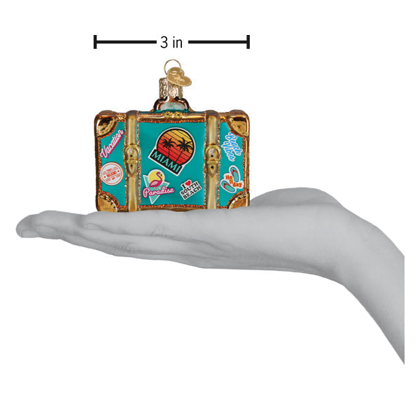 A hand holds a teal Miami Suitcase Old World Christmas Ornament 32659, decorated with travel-themed stickers and a three-inch dimension label.