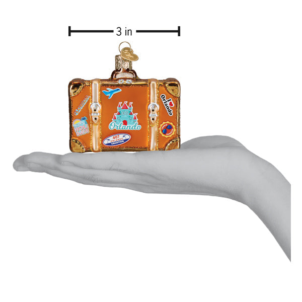 A hand holds an Orlando Suitcase Old World Christmas Ornament 32660, a 3-inch suitcase-shaped decoration featuring travel-themed stickers and an illustration of a castle.