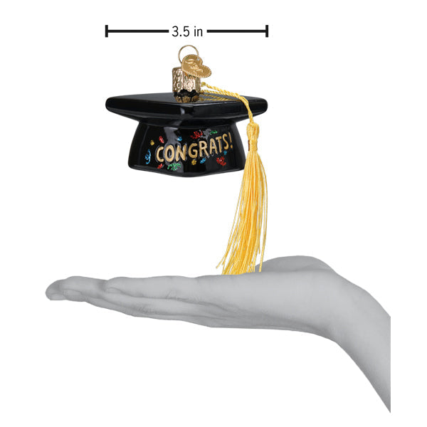 A palm holds a 3.5-inch-wide Graduation Cap Old World Christmas Ornament 36342, featuring a yellow tassel and CONGRATS! written on the front.