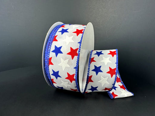 A roll of 1.5X10Y White Blue Red Satin Stars Ribbon (41501-09-25) graces a dark background. The white ribbon features red and blue stars, edged with blue wire, and its loose end partially unravels to reveal the vibrant pattern and silky texture.