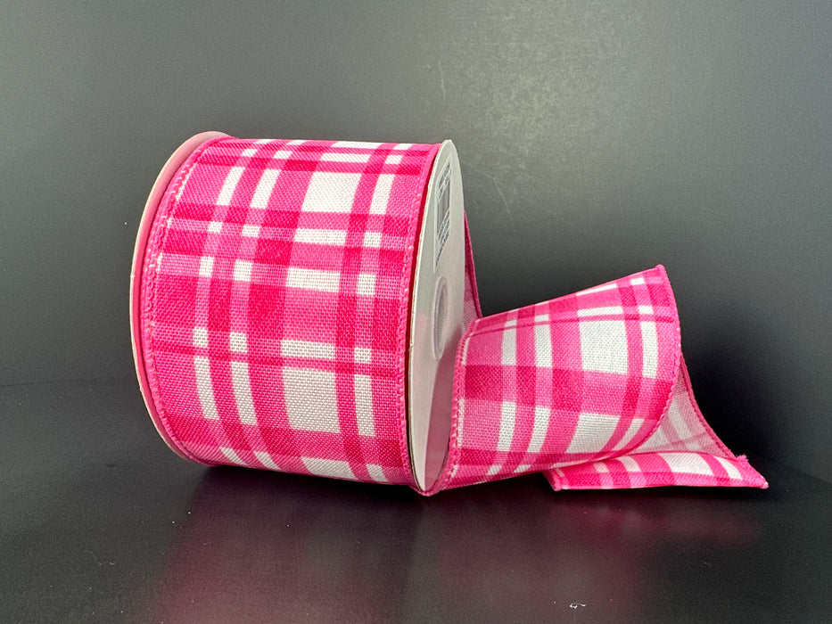 A spool of 2.5"X10Y Pink and White Watercolor Tuttola Plaid Ribbon (41546-40-26) rests on a dark surface, with some of the pink and white ribbon unrolled and folded to the side.