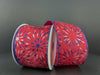 Two rolls of the Red Satin/Red-Blue-Silver Glitter Fireworks Ribbon (2.5X10Y) lie on a gray surface. The edges are elegantly trimmed with blue glitter, while the dark gray backdrop enhances their vibrant hues and starburst pattern.