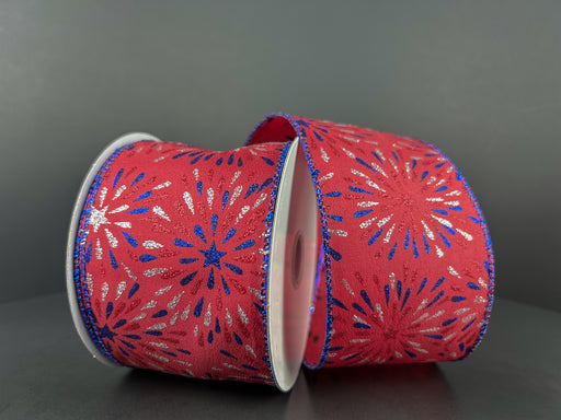 Two rolls of the Red Satin/Red-Blue-Silver Glitter Fireworks Ribbon (2.5X10Y) lie on a gray surface. The edges are elegantly trimmed with blue glitter, while the dark gray backdrop enhances their vibrant hues and starburst pattern.