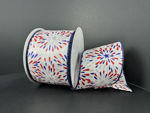 A spool of White Satin/Red-Blue-Silver Glitter Fireworks ribbon (2.5X10Y 42404-40-27) is displayed against a dark background, with its partially unrolled festive design and glitter fireworks motif exuding a celebratory charm.