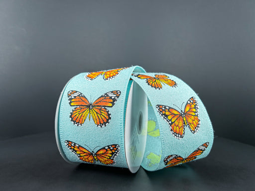 The Monarch Butterfly Ribbon (2.5X10Y Aqua Fuzz Linen/46406-40-45) in vibrant orange and black butterflies on light blue is partially unfurled on a dark gray backdrop.