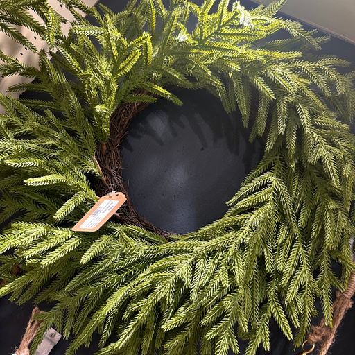 The 24 Evergreen Soft Touch Norfolk Pine Twig Wreath 510700, featuring feathery branches and a small tag, hangs against a dark background with sunlight highlighting the top left.