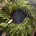 The 24 Evergreen Soft Touch Norfolk Pine Twig Wreath 510700, featuring feathery branches and a small tag, hangs against a dark background with sunlight highlighting the top left.