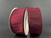 Two spools of 1.5" Burgundy Faux Dupioni Wired Ribbon (10 yards, 72434-09-10), featuring a deep red color and subtle sheen, stand upright against a plain dark background. The ribbon’s elegant wired edges are neatly finished, and the spools have plain silver sides.