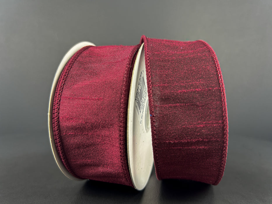 Two spools of 1.5" Burgundy Faux Dupioni Wired Ribbon (10 yards, 72434-09-10), featuring a deep red color and subtle sheen, stand upright against a plain dark background. The ribbon’s elegant wired edges are neatly finished, and the spools have plain silver sides.