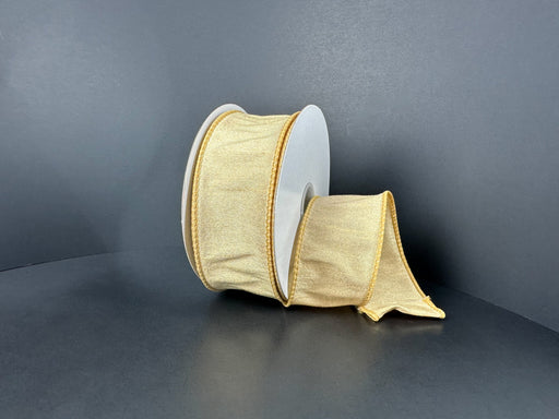 A spool of 1.5X10Y Gold Faux Dupioni Ribbon (72434-09-15) with a subtle sheen rests on a dark surface, part of the elegant ribbon unrolled and lying loosely in front.
