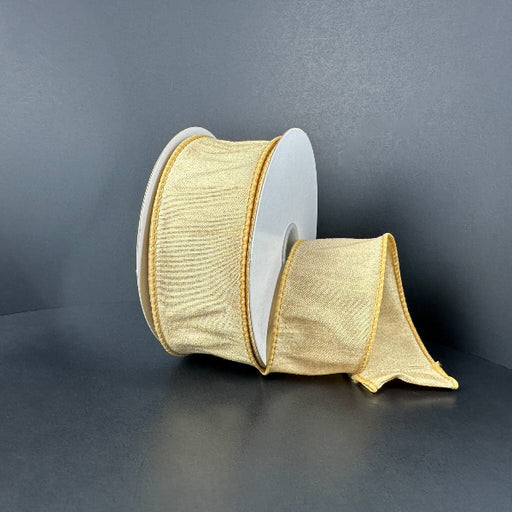 A spool of 1.5X10Y Gold Faux Dupioni Ribbon (72434-09-15) with a subtle sheen rests on a dark surface, part of the elegant ribbon unrolled and lying loosely in front.
