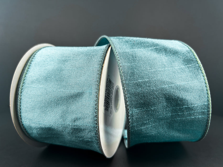 Two spools of 2.5X10Y Dusty Blue Faux Dupioni Ribbon (72434-40-05) sit against a dark background, showcasing their subtle sheen and soft texture.