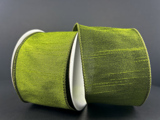 A spool of 2.5" Moss Green Faux Dupioni Wired Ribbon (72434-40-08) with a slightly textured, silk-like sheen unrolls against a dark gray background. The elegant, wide ribbon catches the light beautifully, adding a touch of sophistication to any project.