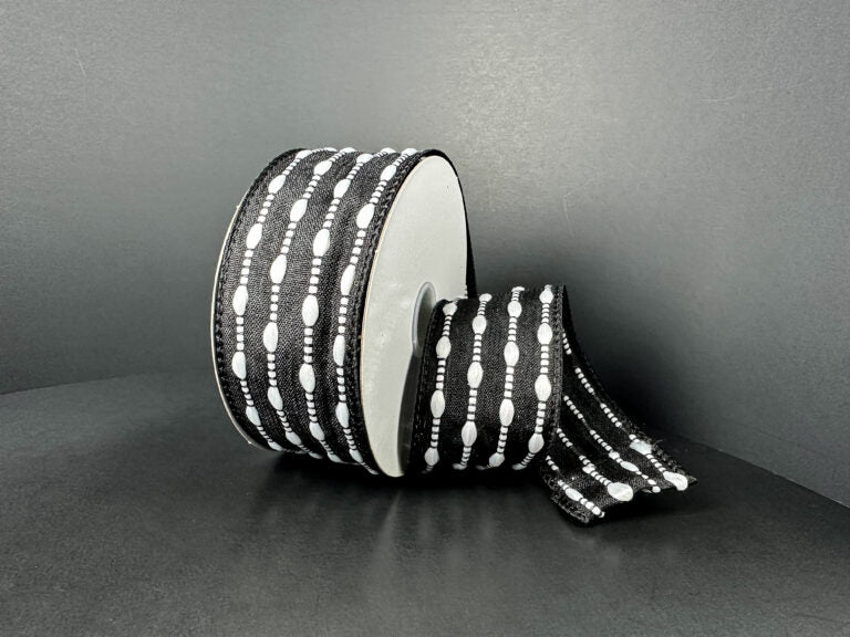 A spool of 1.5X10Y Black/White Snowball Weave Ribbon (73539-09-21), featuring stitched oval shapes and lines in a black and white design on a plain dark background.