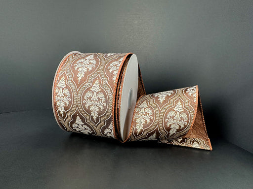 A 2.5x10Y Brown Satin/Demask Quatrefoil Ribbon (73556-40-38) with a white and gold ornate pattern is displayed on a dark surface, partially unrolled and elegantly draped in front.