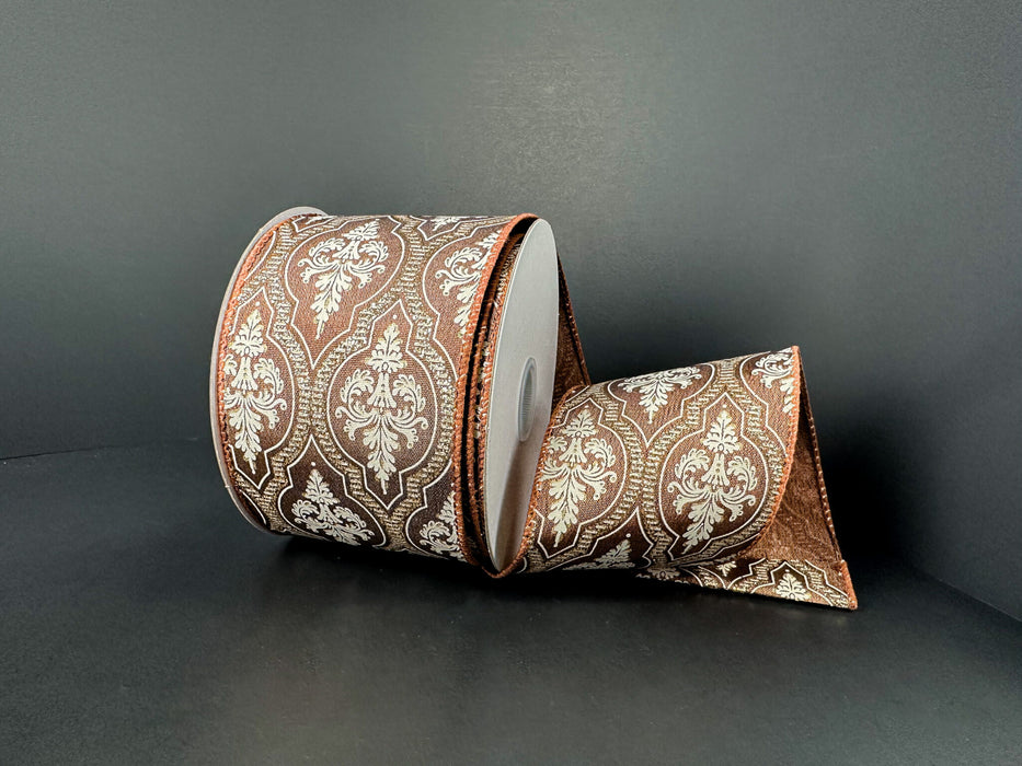 A 2.5x10Y Brown Satin/Demask Quatrefoil Ribbon (73556-40-38) with a white and gold ornate pattern is displayed on a dark surface, partially unrolled and elegantly draped in front.