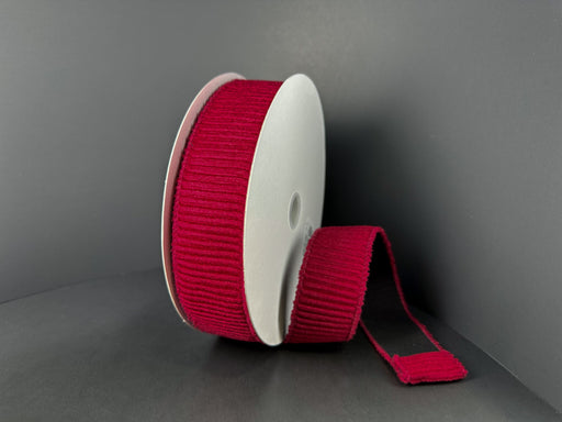 A spool of 1.5x10y Dk Red Ribbed Velvet Ribbon (73561-09-10) stands upright against a dark background, with a length of its deep red ribbon unrolled and lying flat beside it.