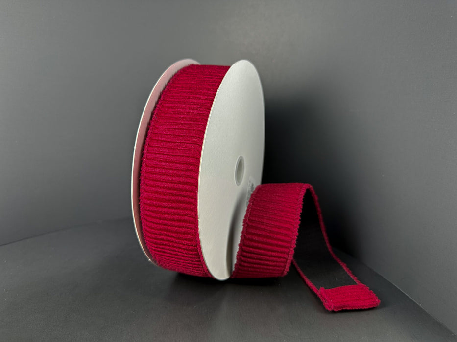 A spool of 1.5x10y Dk Red Ribbed Velvet Ribbon (73561-09-10) stands upright against a dark background, with a length of its deep red ribbon unrolled and lying flat beside it.