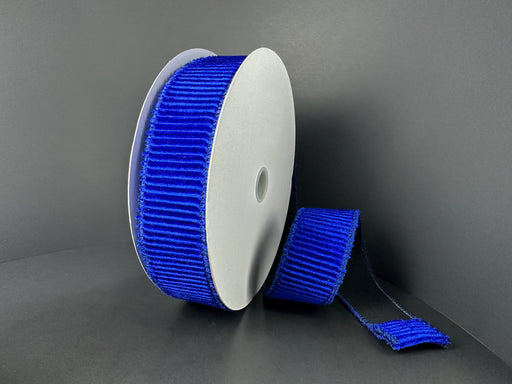 A spool of 1.5X10Y Deep Blue Ribbed Velvet Ribbon (73561-09-25) stands upright against a dark background, with part of the ribbon unrolled and lying flat in front.
