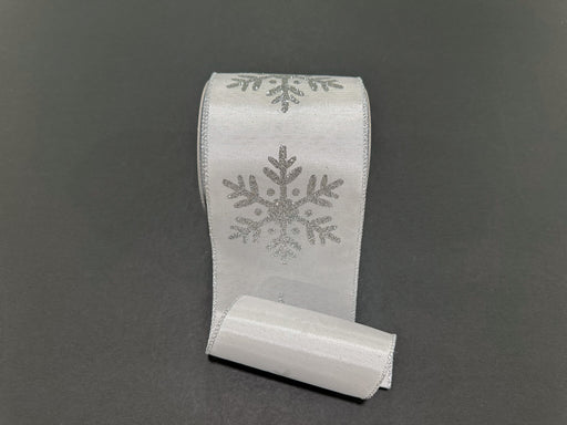 A 4X10Y White Ribbed Satin/Silver Giant Glitter Snowflake Ribbon (73569-04-16) with shimmering snowflake designs lies on a plain dark background.