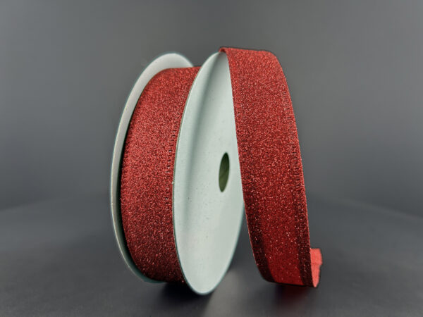 The 7/8"X10Y Red All Flat Glitter Wired Ribbon (76467-05-12) stands upright against a gray background, with some of the red glitter ribbon unrolled and curving gracefully toward the front.