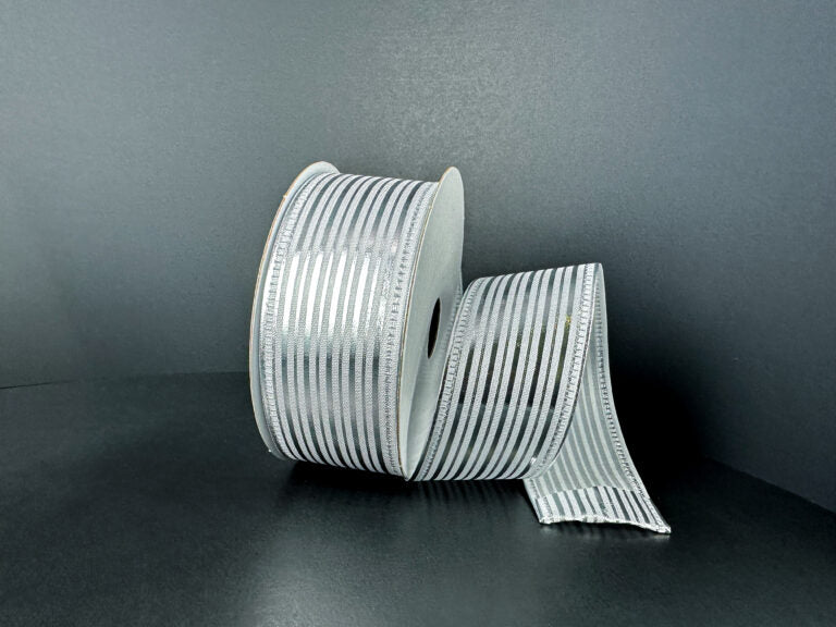 A spool of 1.5X10Y White Satin/Silver Metallic Embossed Vertical Grain Ribbon (76555-09-51) stands upright against a dark background, with a length unrolled and elegantly draped beside it.