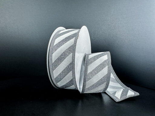 A spool of 1.5X10Y Silver Flat Glitter/White Diagonal Stripes Ribbon (76579-09-01) rests on a dark surface with some ribbon unrolled, showing its shimmering silver and white pattern against a black background.