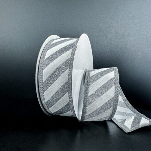 A spool of 1.5X10Y Silver Flat Glitter/White Diagonal Stripes Ribbon (76579-09-01) rests on a dark surface with some ribbon unrolled, showing its shimmering silver and white pattern against a black background.