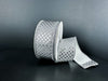 A spool of 1.5X10Y Silver Metallic Dragonscale Dots Ribbon (78521-09-51) rests on a dark surface, its sparkling, diamond-patterned length unrolled and curling forward.