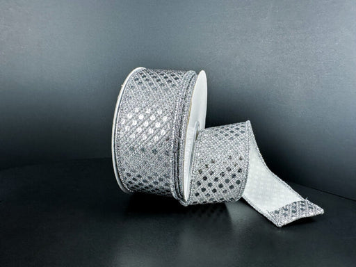 A spool of 1.5X10Y Silver Metallic Dragonscale Dots Ribbon (78521-09-51) rests on a dark surface, its sparkling, diamond-patterned length unrolled and curling forward.